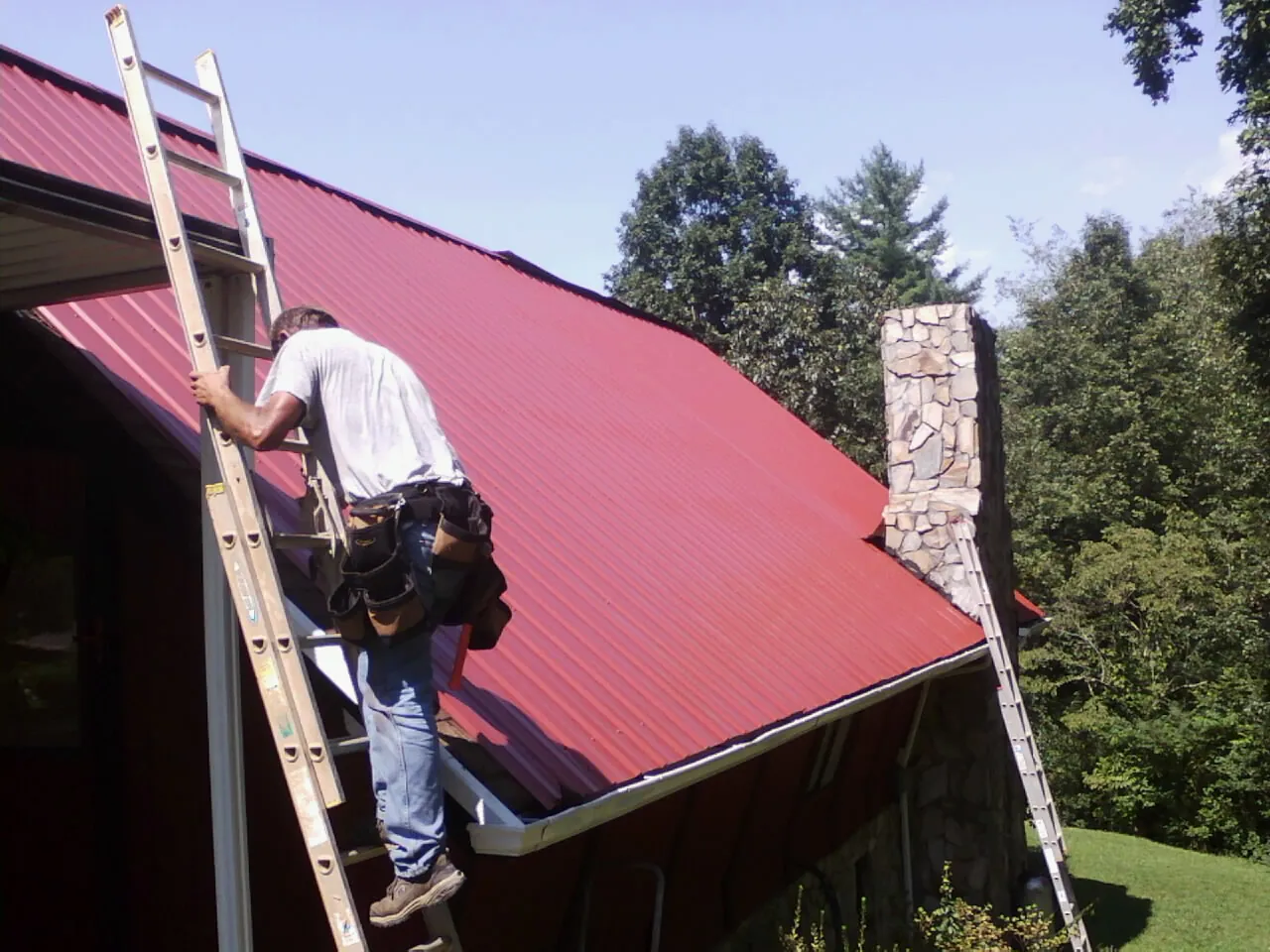 Roofer on a ladder beside a red metal roof and stone chimney — Soffit & Fascia Repair by Roofing Brothers in Shorter